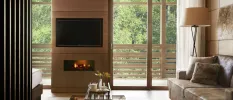 Living area with a plush brown sofa, glass coffee table, wood-panelled wall with a TV and fireplace and full-length glass doors overlooking a forest