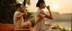 Two people in light, loose clothes sitting in yoga poses doing a breathwork on a wooden deck overlooking a curvy beach and the ocean
