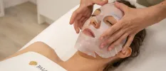 Woman lies with her eyes closed in a white towel as a therapist lies a sheet face mask on her