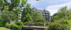 Woman walks over a bridge among gardens in front of a grand multi-storey building under a blue sky
