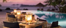 Wooden tables and chairs with black cushions set up for dinner by the poolside, topped with lanterns