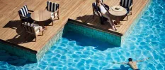 Woman in a white swimsuit and sunglasses relaxes on a blue and white-striped chair on a wooden terrace, as a man floats on his front in the neighbouring pool