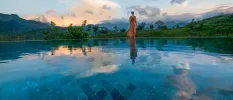 Woman standing in a full-length orange dress in front of a swimming pool overlooking mountains and rainforest as the sky darkens