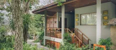 Low-rise villa with pillars, wood panelling, a terracotta tiled roof, gold icons on the wall and a surrounding of tropical greenery