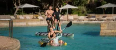 Man, woman and two daughters kayak in a swimming pool surrounded by white parasols and greenery