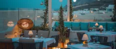 Stylish beach restaurant at night with glass wall surrounds, tables with white tablecloths, and sofas with teal cushions - lighting is soft and in rattan-style lights