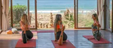 Three women in active wear doing yoga in a studio with a full-length window and a beach and ocean view