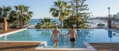 Woman in a striped black and white bikini and man in black trunks walk down steps into a pool set in a wooden terrace, with a view of greenery and the ocean beyond