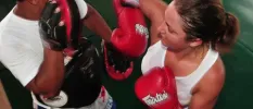 Woman in a white vest and boxing gloves spars with a trainer wearing pads