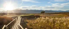 A wooden walkway suspended over green fields and mountains under a blue sky
