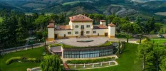 Italian villa with terracotta roof tiles, green window shutters, buttermilk coloured walls and a mustard border, surrounded by green hills and landscaped grounds