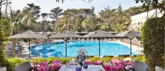 Outdoor swimming pool surrounded by grey parasols and white-cushioned loungers, tropical gardens and a terrace set up for dining with a bed of pink flowers overlooking the pool