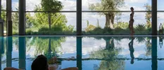 Man relaxing with his hands behind his head in an indoor swimming pool with floor-to-ceiling windows and views of outdoor greenery