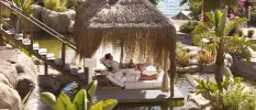Couple in white robes relax on loungers in a thatched-roof cabana surrounded ponds and tropical gardens
