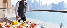 Smiling waiter pours coffee on a balcony with a view of the Dubai skyline, and a table laid with fruit, waffles and juice for breakfast