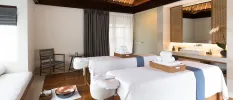 Spa treatment room with two beds covered in white, with rattan trays of white towels on top, and a vanity area with a potted lotus flower