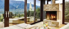 Hotel room with a stone fireplace, dining table and floor-to-ceiling balcony doors showcasing a terrace with loungers and views of the mountains