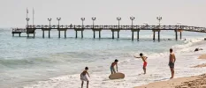 Man and three boys in swimwear play with a surfboard on the shoreline, with a wooden pier in the background