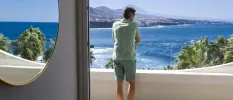 Man in shorts and t shirt looking out at the ocean and mountains from a balcony