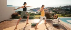 Two women in active wear in warrior pose on a wooden deck next to the pool and a large sound gong