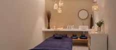 Spa treatment room with cream walls, a massage bed with blue towels, bohemian-style rattan pendant lights and vanity units with flowers and a vase, and a wall-hung mirror