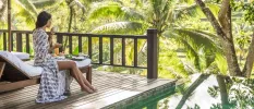 Woman sipping from a white cup with a tray of food in front of her, sitting on a lounger and looking out over a tropical jungle