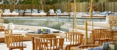 Swimming pool surrounded by wooden fencing, pine trees, white-cushioned loungers and wooden seating