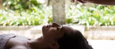 Woman with dark hair and closed eyes lying down surrounded by tropical greenery as a therapist pours oil on her head