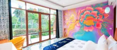 Hotel room with a colourful wall mural in pink, blue and purple, white bedding, a blue velvet throw and wooden full-length patio doors with a view of tropical greenery