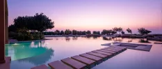 The sun sets over a swimming pool with wooden stepped walkways, sunken seating areas and a fringe of greenery