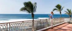 Woman in a blue t shirt looking out at palm trees and the ocean on a sunny paved terrace