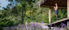 Woman stands in active wear at the edge of a thatched-roof open-sided pavilion with her hands clasped in prayer overlooking tropical greenery