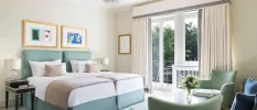Stylish neutral hotel room with twin beds, curtains, teal armchairs and a vase of flowers, and contemporary art hung above the bed