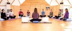 Group sits in a circle in a sheer domed building, while a woman sits at the front of the class next to two white sound healing bowls