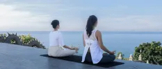 Two women sitting in lotus position on a yoga mat, facing the sea and tropical greenery below