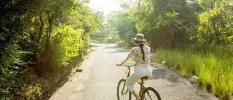 Woman in a hat, white trousers and a yellow floral patterned blouse cycles through a walkway surrounded by greenery