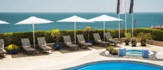Curved swimming pool in a sunny terrace, with white parasols, loungers with blue and white floral cushions, a corner Jacuzzi and views of the ocean