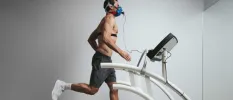 Slim man with dark hair in grey shorts, running on a machine while hooked up to a mask and tubes