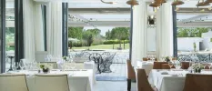 Stylish modern restaurant with white table cloths and curtains, black-framed patio doors, brass pendant lights and an outdoor terrace with black furniture