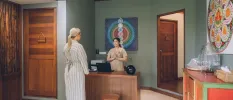 Woman in a striped robe in greeted by a smiling staff member in front of a picture depicting the chakras in a room with wooden doors
