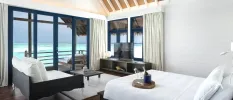 Open-plan villa bedroom and living area with white bedding and sofa, dark-wood furniture and full-length doors with a view of the ocean
