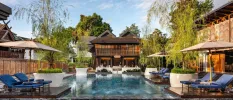 Traditional Thai villa with teak cladding overlooking a tranquil pool, surrounded by blue loungers and white umbrellas under a blue sky 