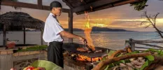Chef in an open-air kitchen cooking over an open flame surrounded by ingredients and overlooking the sea