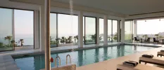 Indoor pool with floor-to-ceiling windows, white-cushioned loungers with striped towels, all overlooking the sea
