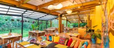Open-air bohemian restaurant with wooden tables adorned with crystals, seating with multi-coloured cushions and a colourful wall mural among yellow walls