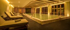 Lantern-lit indoor pool surrounded by cushioned wooden loungers and with wooden beams in the ceiling