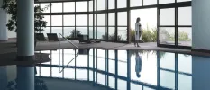 Woman in a black bikini standing next to an indoor pool with floor-to-ceiling windows