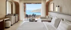 Neutral hotel room with striped bedding, lantern-like wall lights, a wooden dressing table and stall and a furnished balcony with sea and hillside views