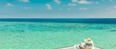 Woman in a straw hat reads a magazine while sitting on a cushion next to the turquoise shallows of the Indian Ocean