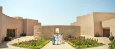 Three women walk through a sunny courtyard surrounded by sand-coloured cube-like buildings, with flower beds leading to a central water feature flanked by stone walls